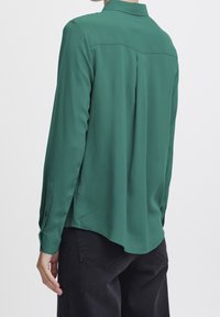 Groene blouse met lange mouwen en kraag, met een rugplooi detail en zijsplitten. Gladde textuur, lichtgewicht stof en met knoopsluiting bij de manchetten.