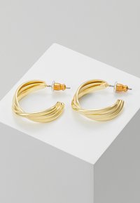 Boucles d'oreilles en métal doré en forme de créoles avec un design torsadé et une finition texturée, maintenues par des fermoirs en argent et en bois.