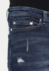 Jeans de mezclilla azul oscuro con una textura desgastada, que presentan un pequeño desgarro y detalles deslavados cerca de los bolsillos y la cinturilla. Acentos de hardware en plata.