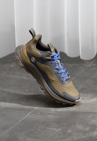 Trail running shoe featuring a khaki mesh upper, dark blue accents, and blue laces. Thick rubber sole with textured grip patterns.