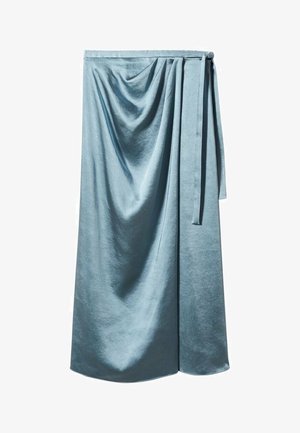 Mujer con un suéter de crochet blanco, falda envolvente de satén azul claro con abertura y sandalias marrones de tacón alto, de pie sobre un suelo liso.