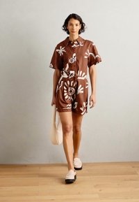 Romper marrone a maniche corte con motivi floreali bianchi, vita legata e bottoni. Il modello porta una borsa leggera e indossa scarpe chiare.