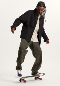 Chaqueta negra abotonada con bolsillos frontales, camiseta beige debajo y pantalones vaqueros verdes olivo sueltos. Patinando sobre una patineta con ruedas rojas.