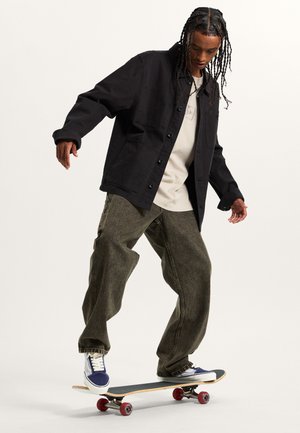 Black button-up jacket with front pockets, beige T-shirt underneath, and loose olive green jeans. Skateboarding on a skateboard with red wheels.