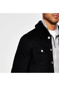 Man som bär en svart denimjacka med svart fluffig krage över en vit och grå mönstrad zip-up skjorta.