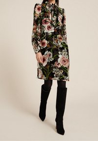 Vestido negro de flores con un cuello alto, mangas largas y vibrantes patrones de rosas y plantas. Combinado con botas negras hasta la rodilla.