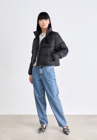 Donna con i capelli neri lisci che indossa un giubbotto invernale nero, jeans azzurri chiari, una collana a catena e scarpe con punta metallica in piedi contro uno sfondo bianco.