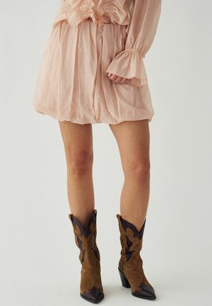 Kobieta ubrana w jasnoróżową mini sukienkę z falbanami oraz brązowe zamszowe kowbojki z ciemnymi skórzanymi detalami, stojąca na jednolitym tle.