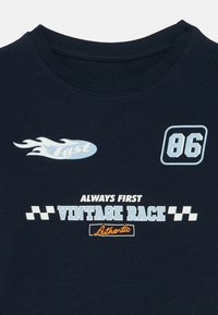 T-shirt azul-marinho com decote redondo, apresentando texto e gráficos de tema de corrida em azul claro e branco: "Rápido," "86," "Sempre Primeiro Corrida Vintage," e "Autêntico."