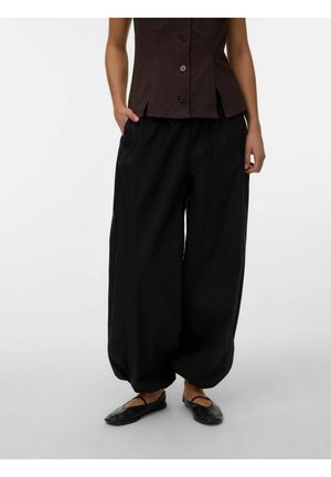 Femme portant un pantalon large noir ample, un haut sans manches marron foncé à boutons, et des ballerines noires Mary Jane avec une bride.
