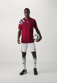 Punane sportlik jersey siniste aktsentidega, valged lühikesed püksid ja mustad jalgpallijalatsid. Hoides musta ja valget jalgpalli, seisab neutraalsel taustal.