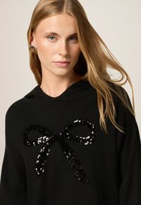 Schwarzer Hoodie mit einem großen Pailletten-Bow-Graphic in verschiedenen Schwarz-Tönen. Glatte Stoffoberfläche, lockere Passform.