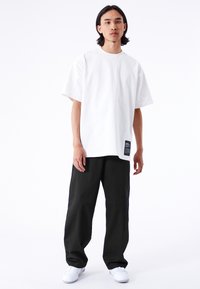 Vit, oversize t-shirt i bomull med rund halsringning, korta ärmar och en svart taggdetalj på nedre vänstra sidan. Kombinerad med lösa svarta byxor.