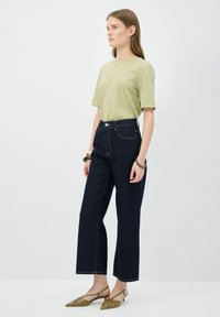 T-shirt verde in cotone con maniche corte, abbinato a jeans a gamba larga in denim scuro a vita alta. Presenta un bottone argentato e cuciture bianche a contrasto.