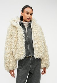 Futrzana kremowa kurtka z kapturem, z zamkiem z przodu i szerokimi rękawami, założona na ciemnoszary strój dżinsowy. Teksturowany, pluszowy materiał.