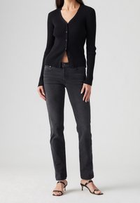 Cardigan negro con canalé, escote en V y botones, combinado con jeans negros de pierna recta y tacones negros con tiras. Textura suave, diseño ajustado.