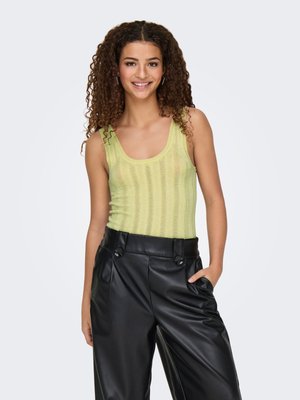 Jeune femme aux cheveux bouclés portant un top en maille sans manches vert clair et un pantalon en cuir noir taille haute, souriante avec une main dans la poche.