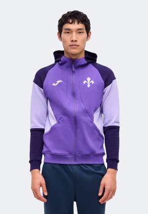 Un homme porte un sweat à capuche de sport zippé violet et lavande, orné de logos blancs, sur un fond uni et clair.