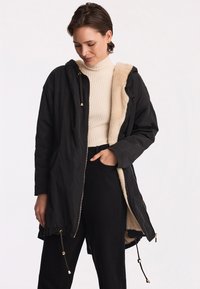 Czarna parka z miękką beżową podszewką z polaru, złotymi elementami ozdobnymi, kapturem i regulowanymi sznurkami u dołu. Nosi się ją na prążkowanej bluzce.