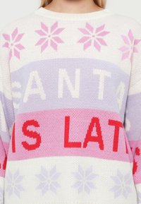 Frau trägt einen weißen Strickpullover mit pinkem und rotem Text sowie Schneeflockenmuster auf der Rückseite.