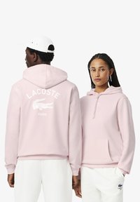 Sudadera rosa claro hecha de tela suave, con un bolsillo frontal y capucha con cordón. En la parte posterior se muestra "LACOSTE" y el logo en blanco.