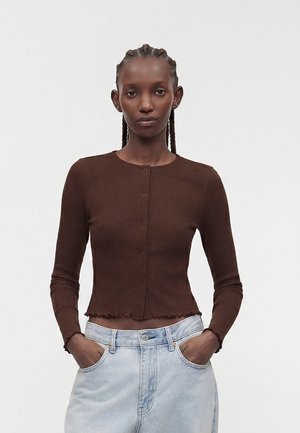 Femme aux cheveux tressés portant un haut marron cintré à manches longues avec boutons et un jean bleu clair, les mains dans les poches, sur un fond uni.