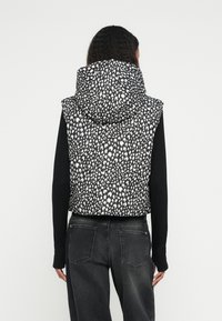 Μπουφάν αμάνικο με μοτίβο σε ασπρόμαυρο, με ψηλό γιακά και κουκούλα, συνδυασμένο με μακρυμάνικη μαύρη μπλούζα και σκούρο γκρι παντελόνι.