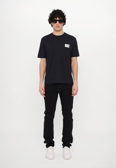 Tricou negru cu mânecă scurtă, având o petică texturată argintie pe partea stângă a pieptului, asortat cu blugi negri și adidași albi.
