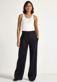 Débardeur blanc avec des bordures noires associé à un pantalon large et sombre. Texture lisse ; pas de motifs visibles. Le mannequin se tient dans un environnement neutre.