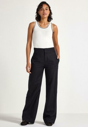 White tank top with black trim paired with dark, wide-leg pants. Smooth texture; no visible patterns. Model stands in a neutral setting.
