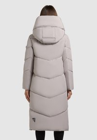 Kvinde iført en lang, lys grå polstret vinterfrakke med en oversized hætte, set fra ryggen mod en ensfarvet baggrund.