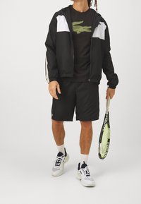 Man i svart och vit sportklädsel som håller en tennisracket, bär sneakers och vita strumpor mot en enfärgad bakgrund.