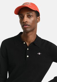 Zwart gebreid poloshirt met een kraag en drie knopen, met een klein embleem op de bovenste linkerkant van de borst. Oranje baseballpet met geborduurde tekst.