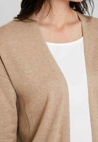 Beige cardigan i mjukt stickat material med djup V-ringning och öppen framdesign, lager-på-lager över en vit blus.