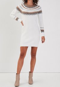 Femme portant une robe-pull en maille blanche avec un motif géométrique marron et noir sur le yoke, associée à des bottines beiges, sur fond uni.