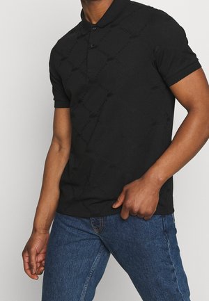 Polo noir à motif texturé, avec patte de boutonnage à deux boutons, manches courtes, porté avec un jean en denim bleu. Finition du tissu lisse.