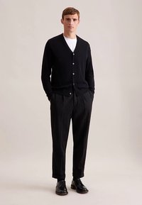 Cardigan noir avec col en V et fermeture par boutons, associé à un pantalon sombre et des chaussures noires, présentant un design simple et des lignes épurées.