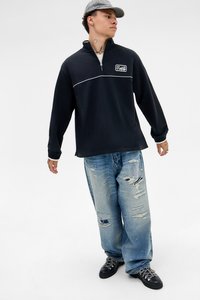 Ung person iført en sort sweatshirt med kvart lynlås, slidte, baggy blå jeans, sorte støvler og en ternet kasket, der står foran en hvid baggrund.