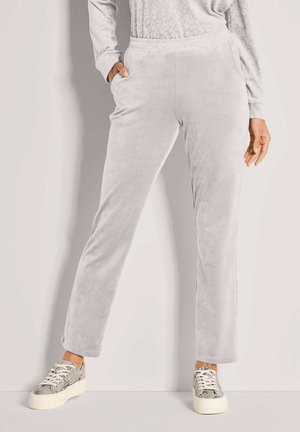 Vrouw in lichtgrijze velours broek en slangenpatroon platform sneakers, staand met één hand in de zak tegen een effen achtergrond.