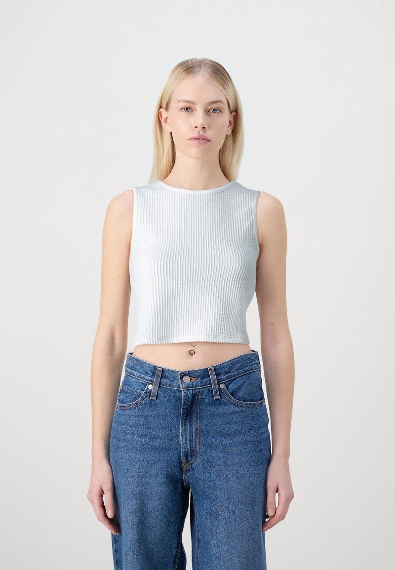 Bílý zkrácený ribbed top vyrobený z texturované tkaniny, kombinovaný s vysokými modrými džíny s knoflíkovým zapínáním a kapsami.