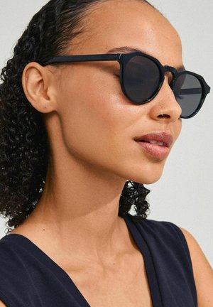 Black round sunglasses with thick frames and dark lenses, featuring a geometric design and a matte finish. Model has curly hair.