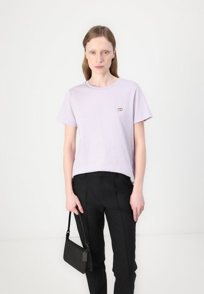 T-shirt en coton lilas avec un col rond et des manches courtes, arborant un petit logo doré. Associé à un pantalon noir sur mesure et un sac à main noir.