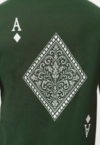 Σκούρο πράσινο ύφασμα με μεγάλο λευκό περίτεχνο μοτίβο σε σχήμα ρόμβου και δύο μικρότερα σύμβολα "Α" και ρόμβου, που μοιάζουν με την Άσσο Καρό από μια τράπουλα.