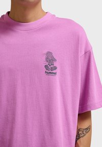 Mężczyzna w różowej koszulce z małą grafiką biegającej rękawicy bokserskiej oraz napisem „hummel sportswear” na piersi.