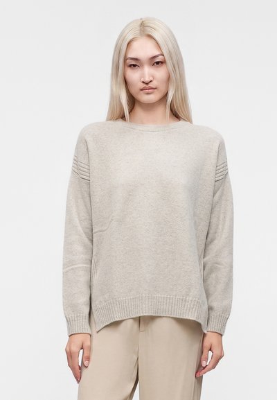 Femme aux longs cheveux blonds portant un pull en maille beige clair et un pantalon beige, debout devant un fond blanc uni.