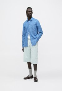 Jasnoniebieska koszula w paski zapinana na guziki, jasne krótkie spodenki dżinsowe, szare skarpetki oraz ciemne mokasyny. Model stoi na jednolitym białym tle.