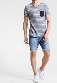 Pruhované tričko s námořnickou kapsou, spárované s vyhrnutými denimovými šortkami. Model má na sobě bílé tenisky. Neformální letní outfit.