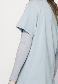 T-shirt oversize azzurro chiaro con maniche corte, indossato sopra una maglietta attillata grigia a maniche lunghe, che mette in evidenza trame morbide e un design informale.