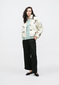 Giacca bianca pelosa con ricamo floreale, maglione azzurro chiaro sotto, pantaloni neri a gamba larga e scarpe nere piatte.