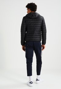 Schwarze Pufferjacke mit Kapuze, horizontale Steppung, kombiniert mit marineblauen Jogginghosen mit Seitestreifen. Model steht mit dem Rücken zur Kamera, vor einem schlichten Hintergrund.